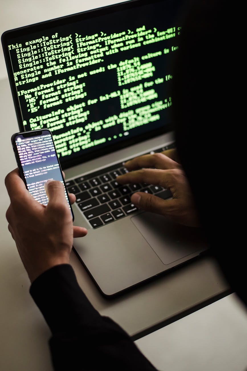 unrecognizable hacker with smartphone typing on laptop at desk Why Email Security Still Gets Overlooked (And What To Fix Today)