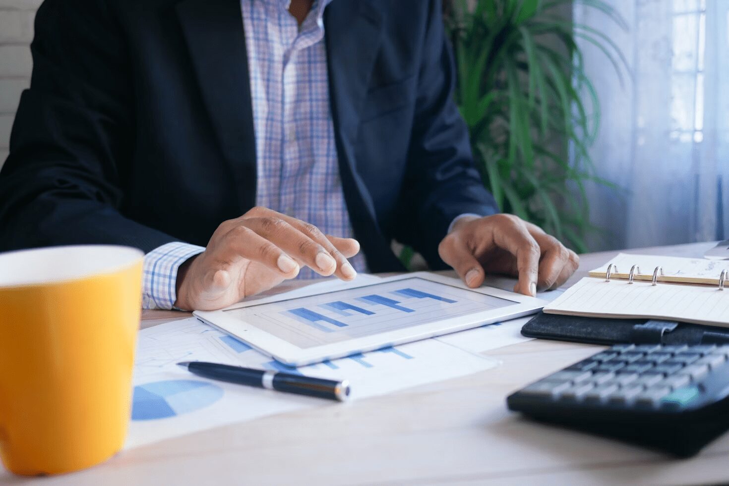 A person in a suit using a tablet to analyze financial data, with a coffee cup and notebook on a desk.