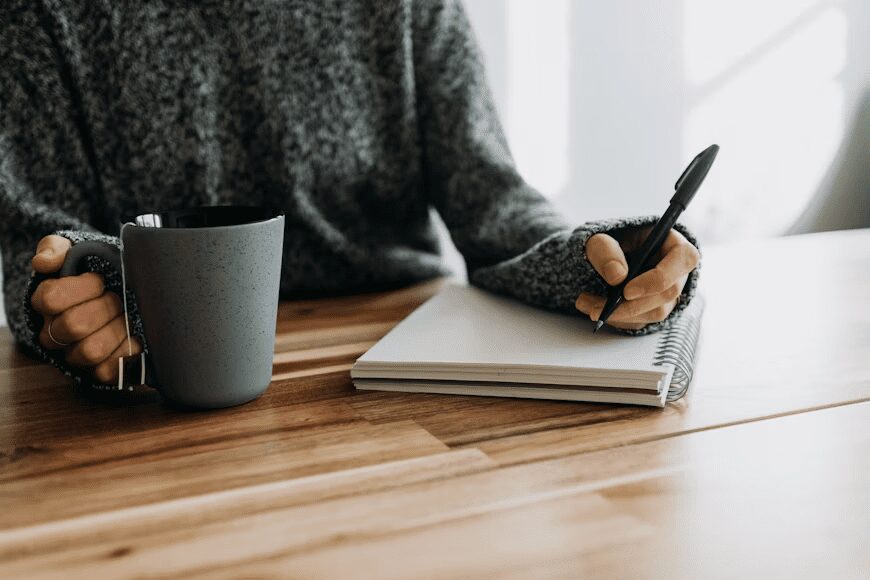 A person wearing a cozy sweater holding a gray mug in one hand and writing in a notebook with a pen on a wooden table. 10 Steps to Build a Successful Writing Business
