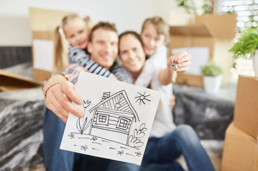 A happy family with two children is sitting in their new living room surrounded by moving boxes. The father holds up a drawing of a house while the mother shows a set of keys. Is Building Your Own Home the Ultimate Dream or a Costly Mistake?