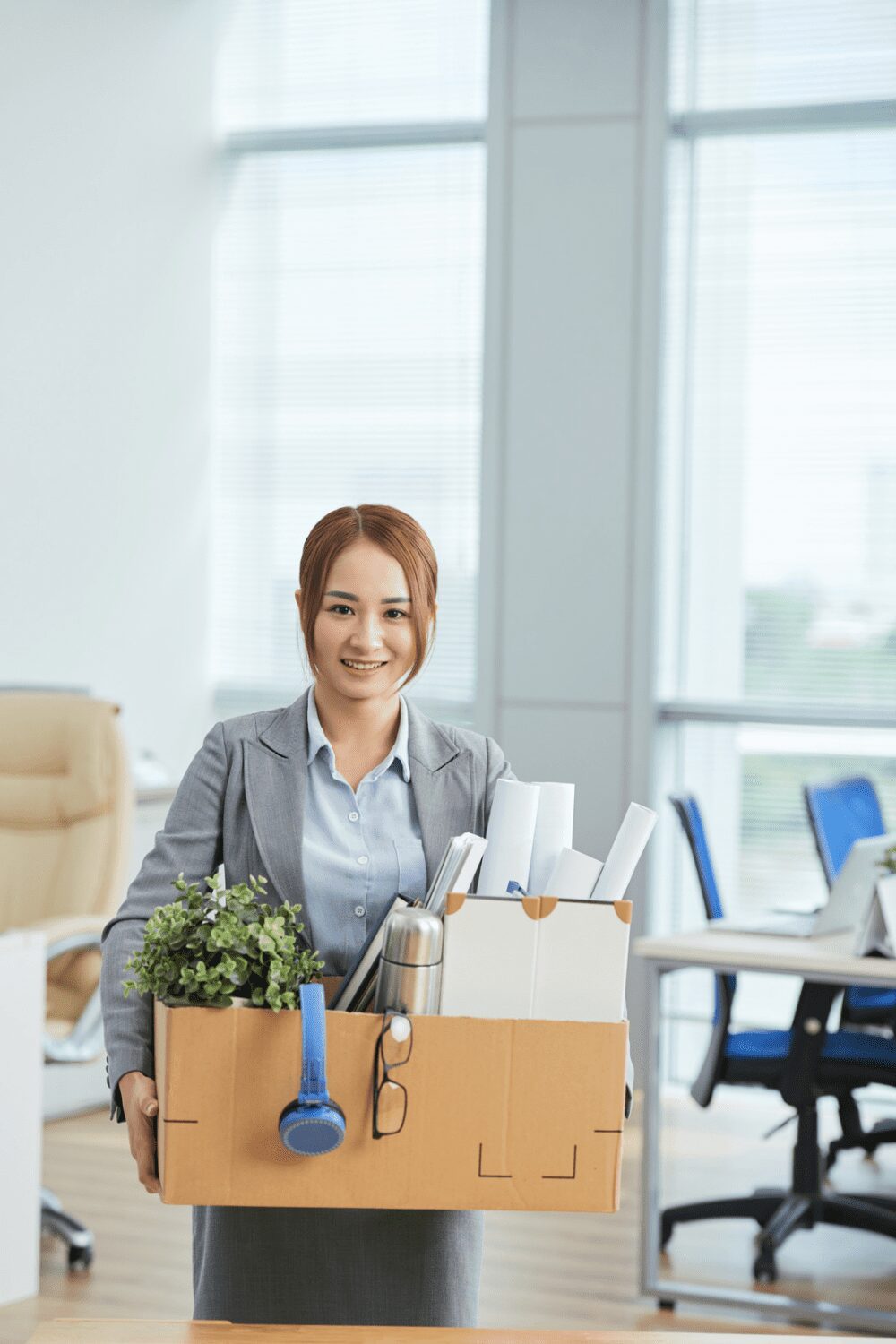 A woman in a business suit is smiling while carrying a cardboard box filled with office supplies, a small plant, a thermos, and some documents in a modern office setting. Relocating for Work: 6 Smart Strategies for a Smooth Move