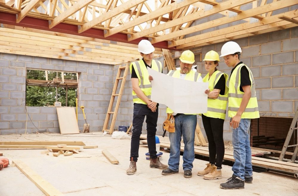 A group of construction workers examining blueprints at a building site, surrounded by wooden beams and unfinished walls. Is Building Your Own Home the Ultimate Dream or a Costly Mistake?