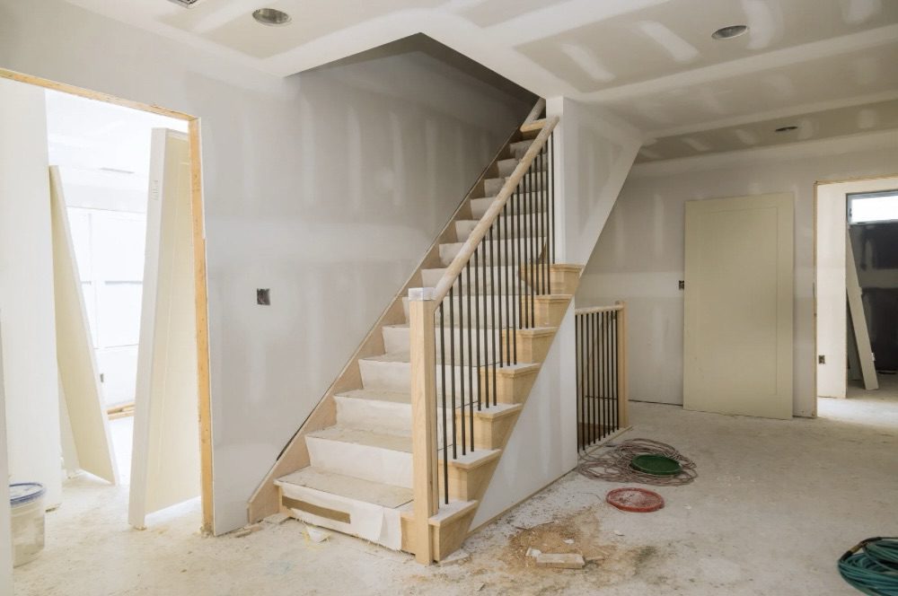 Interior view of a partially constructed home featuring a wooden staircase, with unfinished walls and materials scattered on the floor. Is Building Your Own Home the Ultimate Dream or a Costly Mistake?