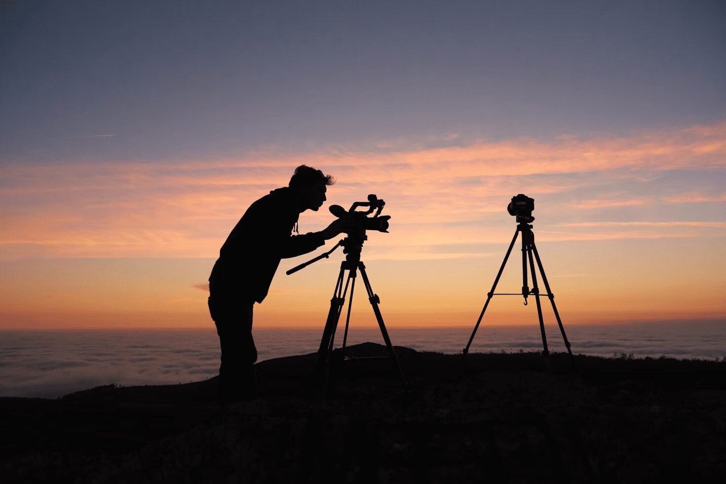 Silhouette of a person adjusting a camera on a tripod during sunset, with another tripod in the background. 6 Careers That Let You Travel and Work At The Same Time