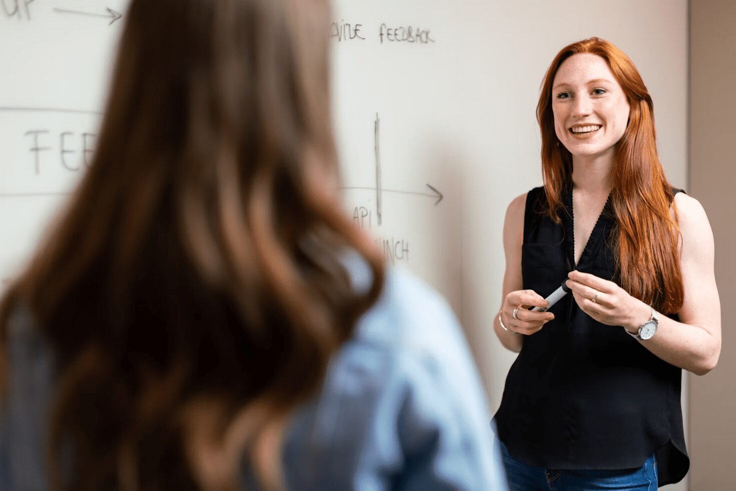 A woman with long red hair and a black sleeveless top smiles and engages with another person in a professional setting, as she holds a marker and stands in front of a whiteboard. 6 Careers That Let You Travel and Work At The Same Time