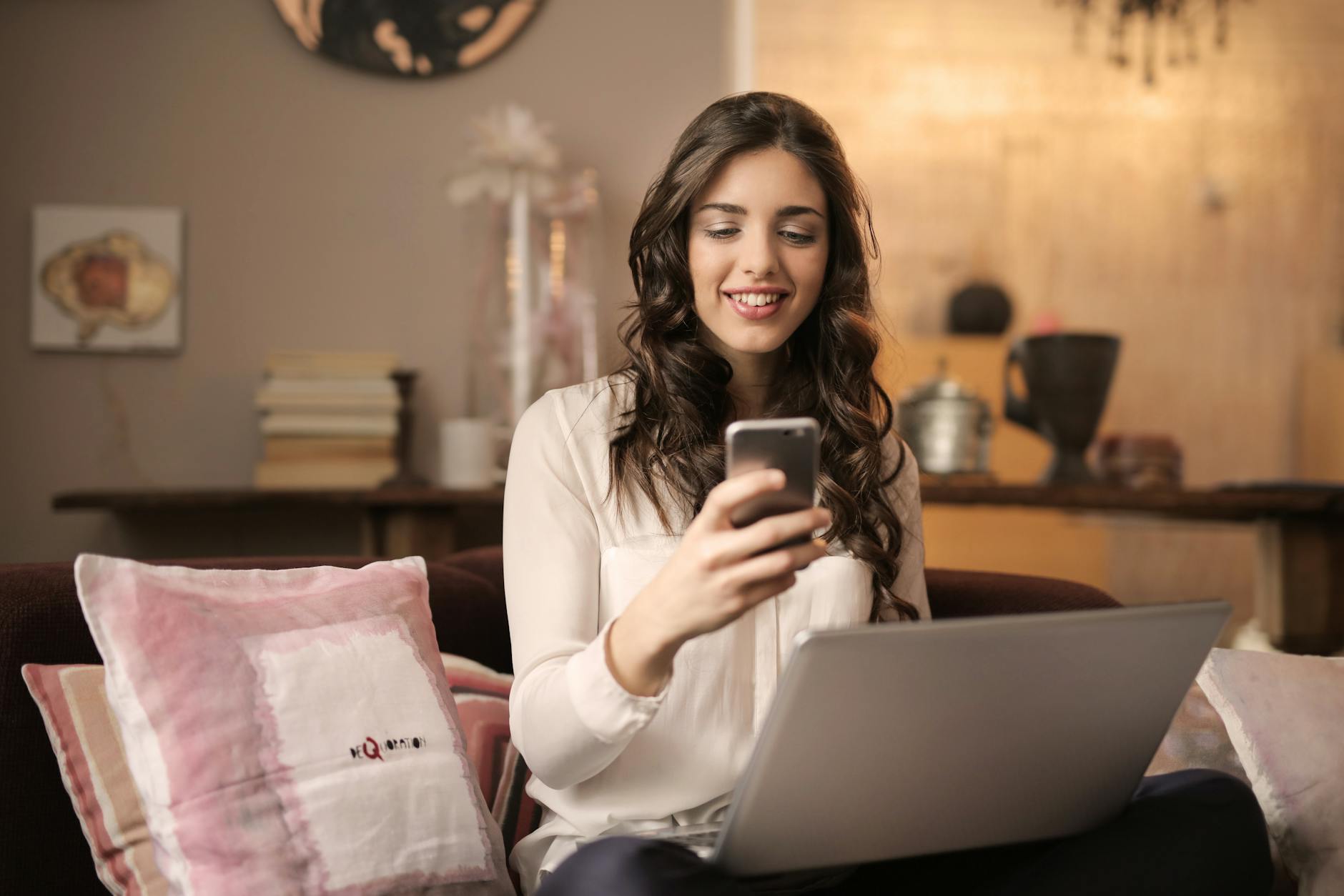 woman sitting on sofa while looking at phone with laptop on lap The Sanity-Saving Guide for Online Store Owners